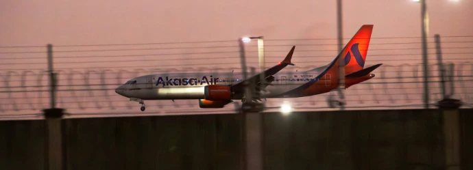 An Akasa Air passenger jet landing behind a wire fence at low light